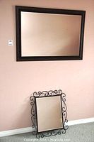 Photo showing both mirrors, the larger wood framed bevelled glass mirror hanging on the wall above, and the smaller decorative wrought iron framed mirror resting on the floor against the wall