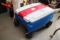 Closed Rubbermaid rolling cooler with blue base, white lid and red latch, showing wheels and handle for rolling.