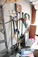 Wide view of garden tools including rakes, shovels, broom, garden baskets hanging on wall, with garden hose and small stool nearby