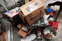 Various boxes and items including a metal bucket with rope and wooden duck decoys, red gas cans, and a box filled with hand tools and hardware supplies scattered on a floor.