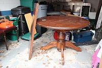 Round wooden pedestal dining table with one leaf leaning beside it. Table surface shows wear and scratches. The table has a single pedestal base with three curved legs.