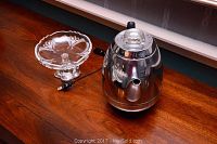 Photo of chrome General Electric electric coffee percolator with black handle, electrical cord, and clear glass bowl on wooden surface.