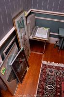 Corner of a room with four framed works leaning on the wall and one on a chair, showing various art prints and a photo.