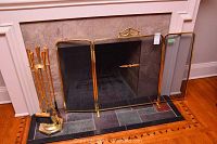 Full view of brass framed fireplace grate with three screen panels, brass fireplace tools, and brass log holder on tiled hearth.