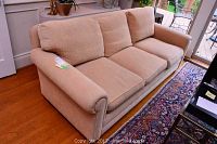 View of the entire beige fabric sofa couch, showing three seat cushions and three back cushions, placed on wooden floor near a window and a rug.