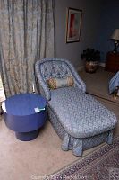 Chaise lounge upholstered in blue-patterned fabric with matching decorative neck roll pillow beside a blue mid-century round side table.