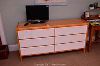 Front view of wooden dresser with light wood body and white drawer fronts, TV and remote on top.