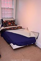 Single white bed frame with mattress, floral duvet, and linens in a corner of a room by a window.