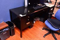 Black wooden office desk with contents such as computer keyboard, mouse and speakers on top, blue desk chair beside it, showing general desk shape and context in room.