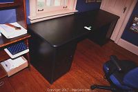 Full view of the black office desk showing the top surface, drawers, and legs in a room with wooden floors.