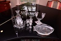 Glassware set arranged on black table including various vases, sherry glasses, double candle stick holder, and fish dish
