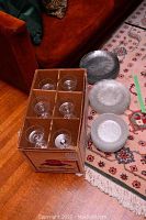 Box containing 12 clear glass sherbet glasses with stems and three different sizes of clear glass plates placed on a rug.
