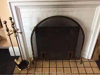 Fireplace with black metal mesh screen in front, arched top, and brass tool rack to left side with four hooks and round base.