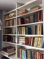 Wide view of multiple white shelves filled with assorted books of various sizes and colors, some stacked horizontally and vertically.