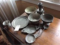 Photo showing the entire set of 11 pewter items arranged on a wooden table including bowls, plates, cup, ladles, pans, and pitcher.