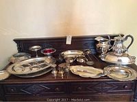 Photo showing full assortment of silver plated items arranged on wooden dresser including coffee pot, pitcher, trays, and smaller serving ware.