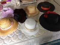 Overview of all six hats on mattress including straw, fur and fabric materials with various decorative ribbons and bands.