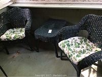 Full set view including two black wicker chairs with white and green floral cushions and matching wicker table on floor.