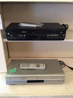 Front view of the black Toshiba DVD player with remote control on top, placed on a shelf with white wall behind.