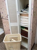 White shelving unit with various folded towels in white, cream, beige, yellow-green, and brown colors, hair dryers on top shelf, and plastic beige laundry hamper with ventilated sides and a lid inside it.