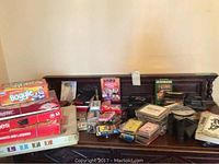 Photo showing a spread of board games, card decks, CDs, binoculars, cassette tapes, and other small items resting on a wooden surface against a beige wall.