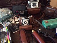 Collection of vintage cameras and accessories showing Kodak, Pentax cameras with leather cases and original film boxes.