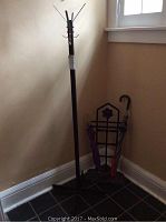 Full view of tall coat rack standing next to black metal umbrella stand holding several umbrellas in a corner by a window.