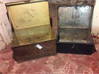 Two metal trunks, one larger bronze-colored and one smaller black, both open and showing interiors with wear and dirt.