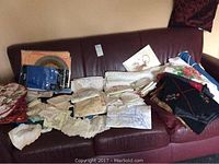 Photo showing a collection of assorted table linens laid out on a dark sofa, including folded embroidered tablecloths, lace napkins, and placemats with floral designs.
