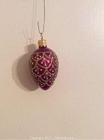 Photo of a fuschia egg-shaped ornament with gold and silver glitter designs hanging on a thin wire against a plain wall.