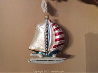 Front view of the hand blown and hand painted glass catamaran boat ornament with red and silver sails hanging on a wall.