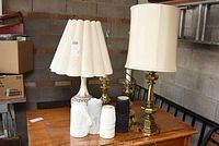 Overview of four lamps on wooden table including glass and metal lamp with large fabric shade, brass lamps and various mini lamp shades.
