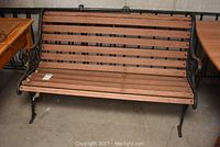 Front view of a wooden slat bench with wrought iron frame and floral armrest design, placed indoors on concrete floor.