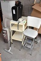 Photo showing vintage stool, white rotary phone on cream metal cart, black paper shredder, and Electrolux vacuum with hose