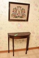 Photo showing wood demi lune table with semi-circular top and slender legs, positioned below the framed needlepoint on floral wallpaper.