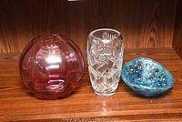 Three glass pieces on wooden shelf: cranberry glass vase, clear cut glass vase, and blue blown glass bowl.