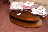 Side and top view of the oval coffee table made of thick glass top supported on a wooden base with metal rods. Wooden lower shelf visible. Light scratches on wood visible.