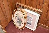 Five framed pieces of art leaning against wood panel wall, view of oval floral prints with gold frames and the rest behind