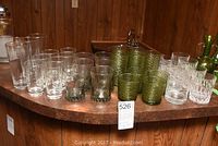 Wide shot showing multiple sets of vintage bar glasses on a curved counter, including tall clear glasses, green textured tumblers, and other clear textured glasses.