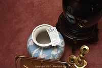 Photo shows a blue ceramic vase with floral and bird motifs, positioned near a black ceramic vase on a wooden stand, brass candle sticks, and a brass decor piece.