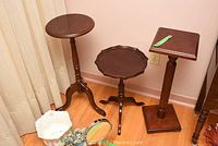 Three wooden plant stands arranged in a corner. Left stand has a round top, middle stand has a scalloped top, right stand has a square top.
