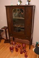 Front view of vintage wooden china cabinet with carved details, glass door, and drawer at bottom. Cabinet stands on four legs on hardwood floor, with several red glass decorative items placed in front.