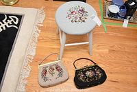 Overview showing round stool with floral needlepoint upholstery and two needlepoint purses on wooden floor.