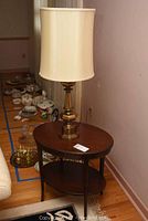 Overall view of the oval wooden side table with brass table lamp on top, showing the setting in the room and some additional items in the background.