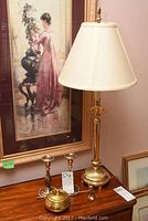 Brass table lamp on wood table next to framed print and other brass and metal decor items