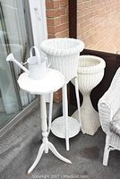 Photo showing two white wicker plant stands, white wooden side table, and white watering can near a brick wall and patio door.