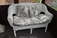 Front view of white wicker loveseat with two floral seat cushions and two floral backrest cushions showing curved arms and woven wicker texture.