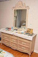 Full view of the dresser and mirror set showing the beige distressed dresser with six drawers and white ornate handles, topped with various items on paper, and the mirror mounted above it on the wall.