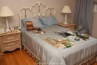 Full view of the double bed with metal and brass painted headboard, blue linens, nightstands with lamps, and various jewelry items displayed on the bed.
