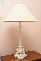 Pair of matching white table lamps on brown wooden surfaces against a pale pink wall, showing front and side views with wide fabric shades and detailed carved bases.